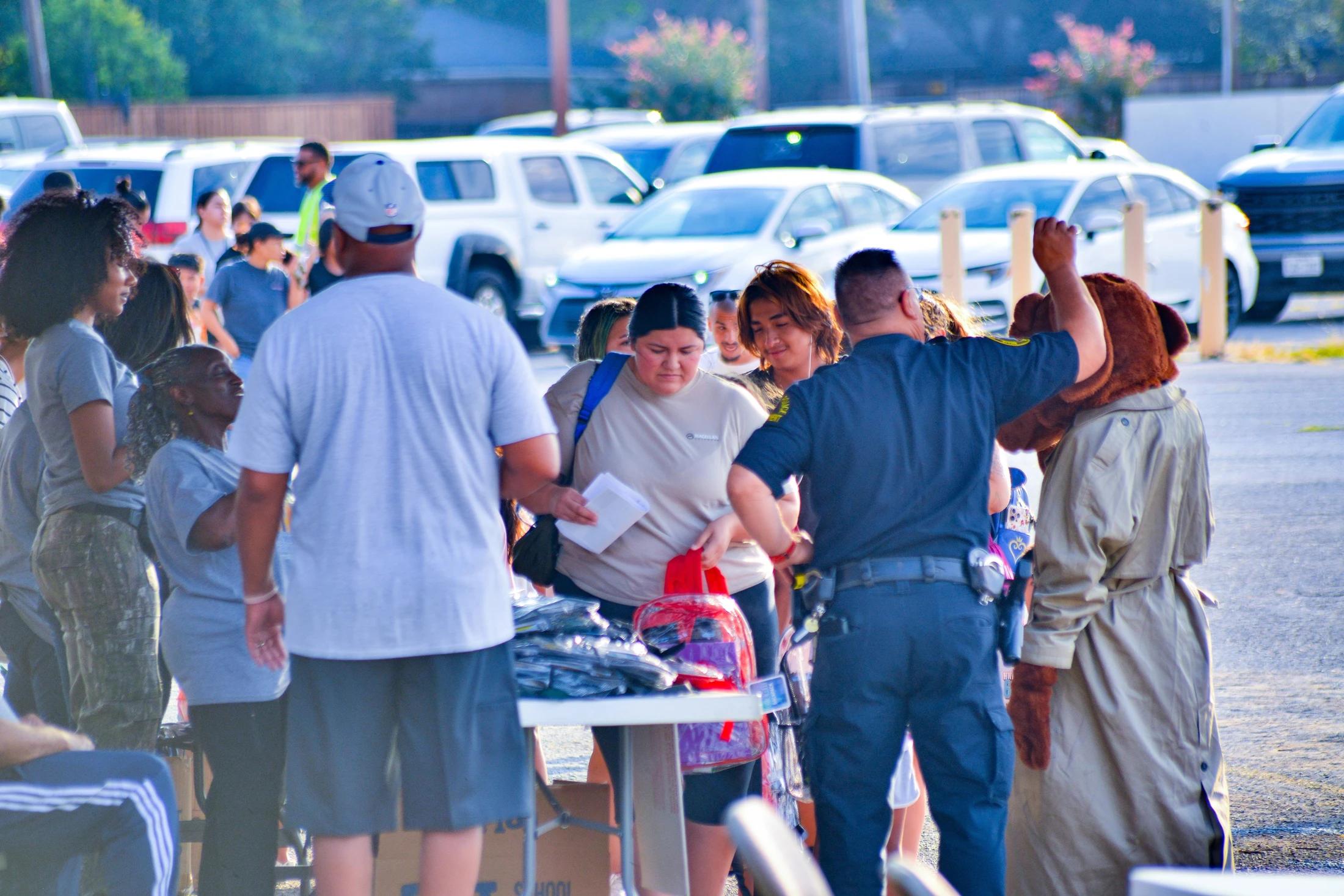 A police officer and a person in a mascot costume interact with a crowd of people at an outdoor community giveaway event in a parking lot.