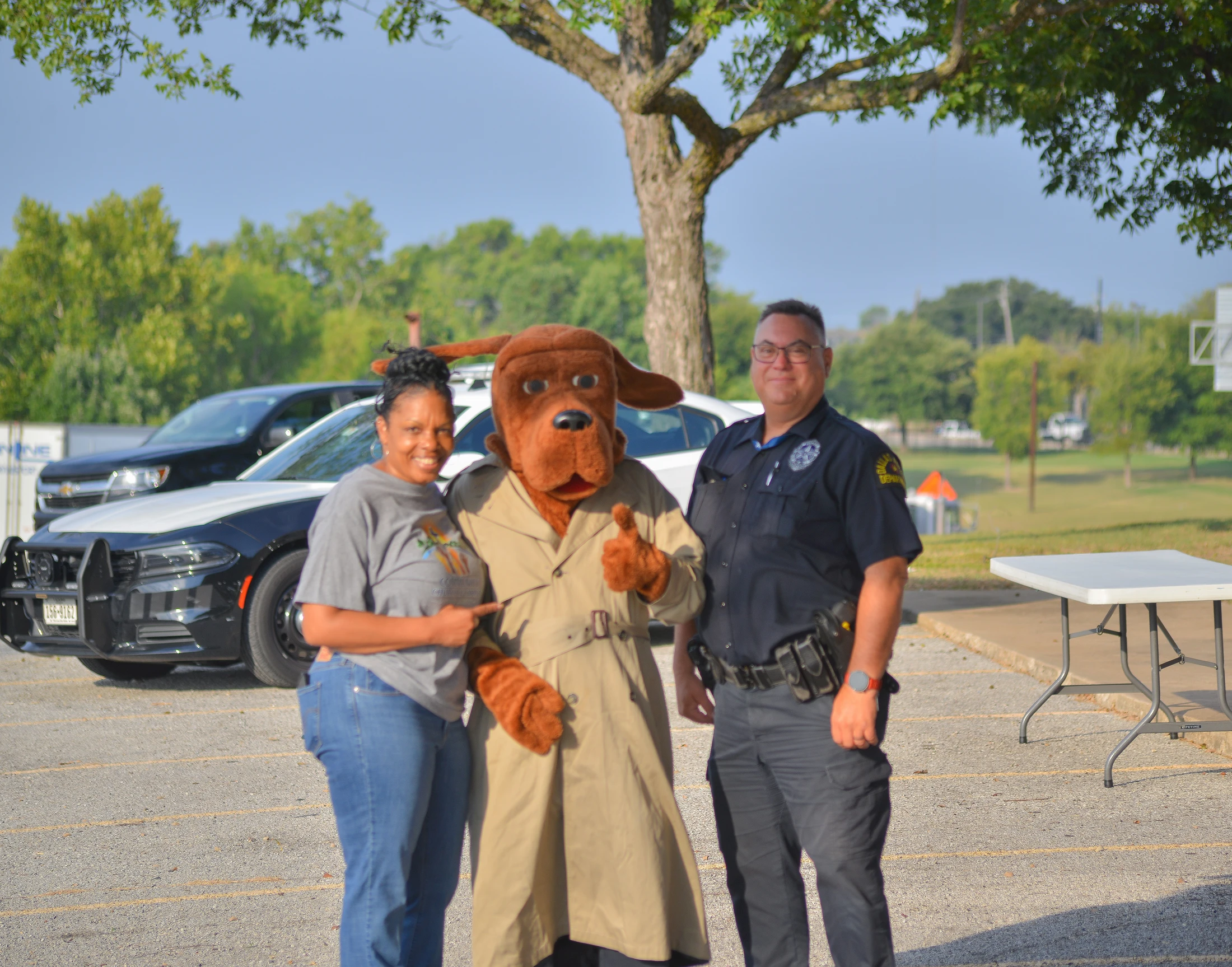 A woman, a police officer, and a person in a McGruff the Crime Dog mascot costume smile for a photo outdoors in a parking lot with a police car behind them.