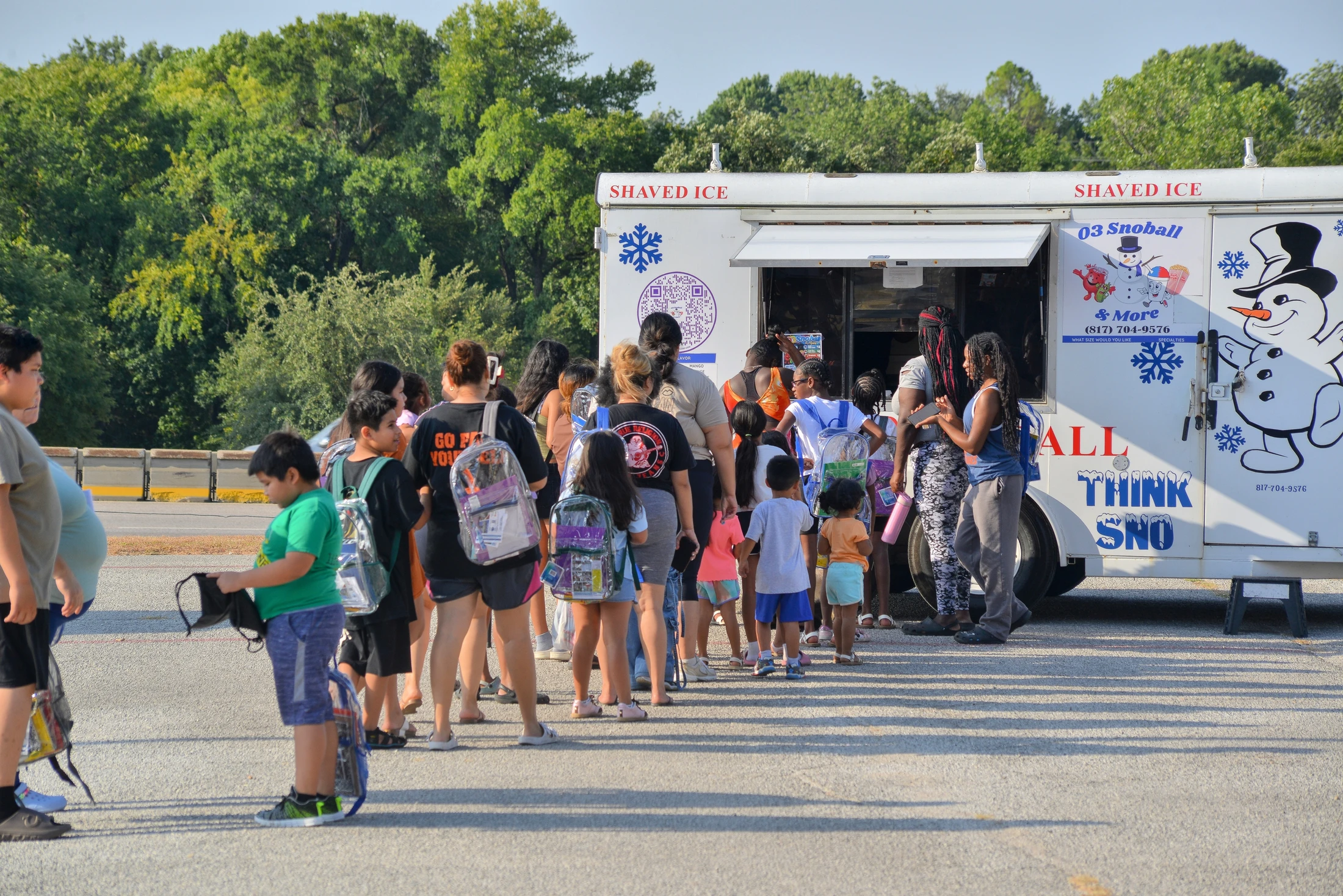 A diverse group of children and adults standing in line at a white shaved ice food truck in a parking lot on a sunny day.