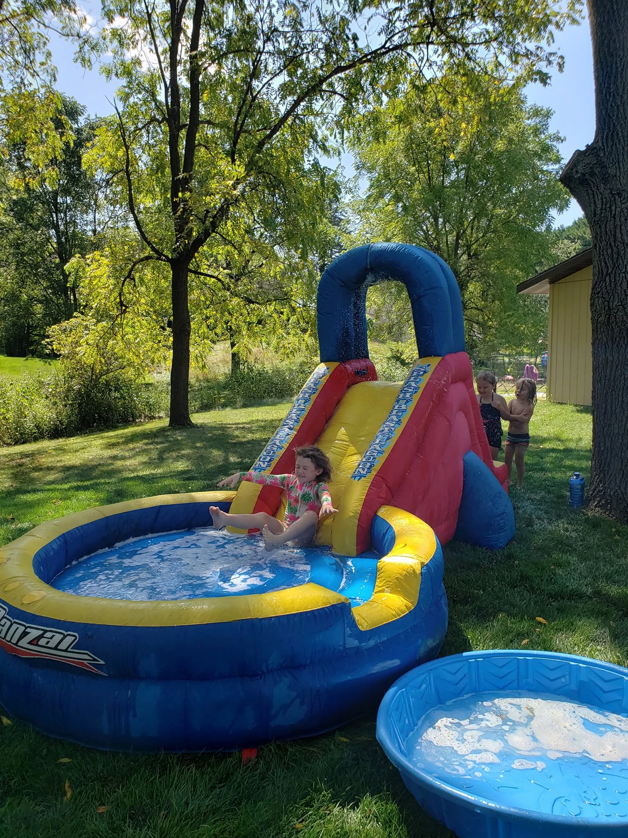 Three young children play on a large, colorful inflatable water slide in a grassy backyard on a sunny day. One child splashes into the attached pool.