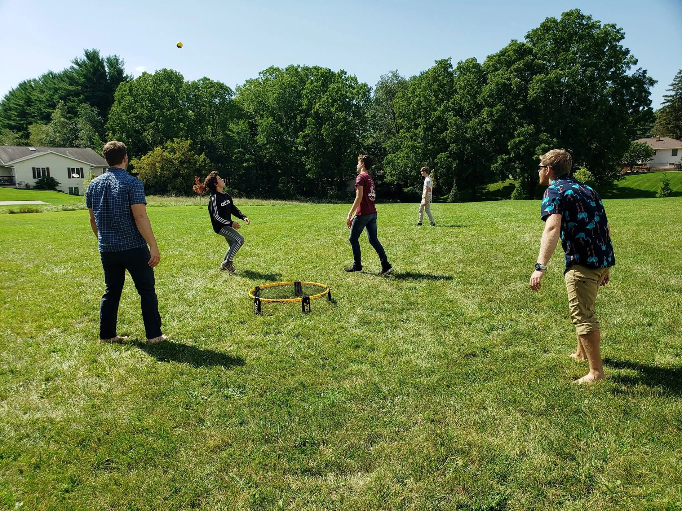 A group of five friends play a game of Spikeball on a large green lawn on a sunny day, with a yellow ball in the air and trees in the background.