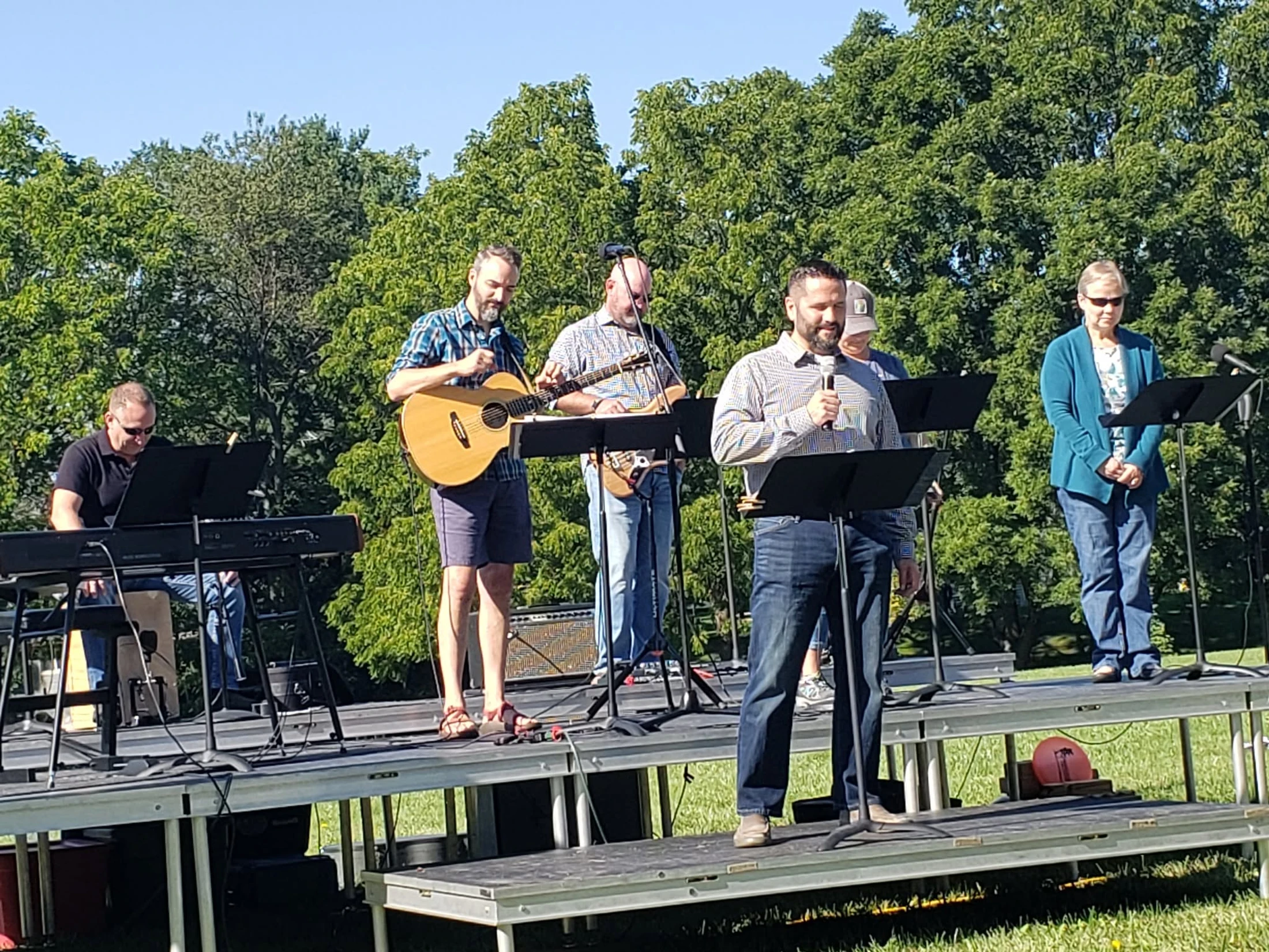 A band of five people performs on a temporary stage in a park. Musicians play keyboard and guitars while a man at the front speaks into a microphone.