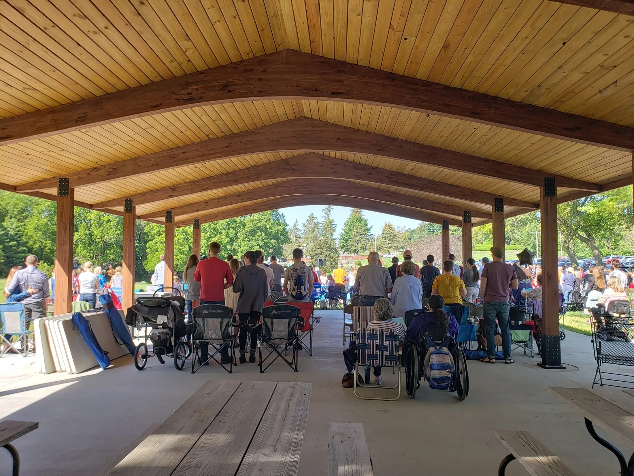 A large crowd of people gathered under a spacious wooden park pavilion on a sunny day. Many are standing or sitting in camp chairs, facing an event.