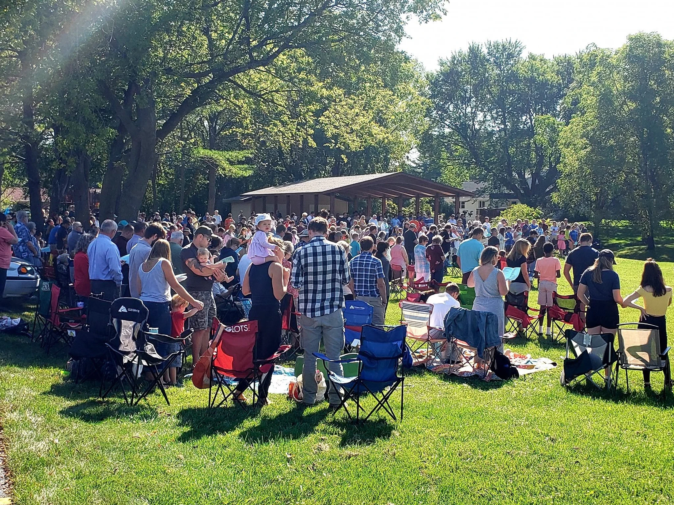 A large crowd of people gathered on a sunny day in a park, sitting on blankets and lawn chairs facing a pavilion surrounded by lush green trees.