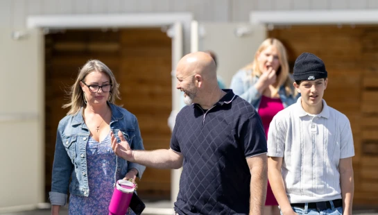 A group of four people walking on a wooden patio. A man in a polo shirt gestures as he talks to a woman in a floral dress, next to a teen in a beanie.