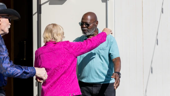 A woman in a bright pink jacket greets a man in a blue shirt outside a building, while another man in a cowboy hat looks on from a doorway.