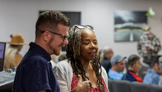 A man with glasses smiles while listening to a woman with long dreadlocks who is speaking and gesturing with her thumb up in a room with rows of chairs.