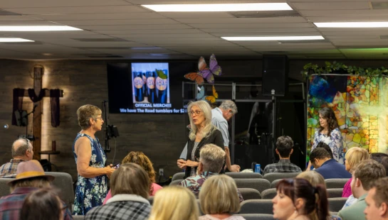 An audience sits in a church hall while two women stand near the front talking. A screen in the background advertises official merchandise.