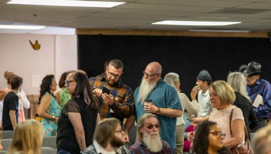 A diverse group of people mingling in a room. Two men stand in the center, one showing the other his phone, while others talk and sit in the background.
