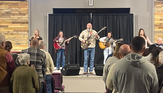 A band with several guitarists and singers performs on a stage for a congregation. The audience is standing and facing the stage in a modern church.