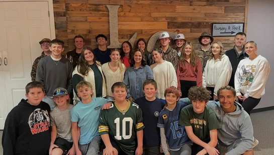 A large group of smiling teenagers and young adults pose for a photo indoors against a rustic wooden wall with a large letter L.