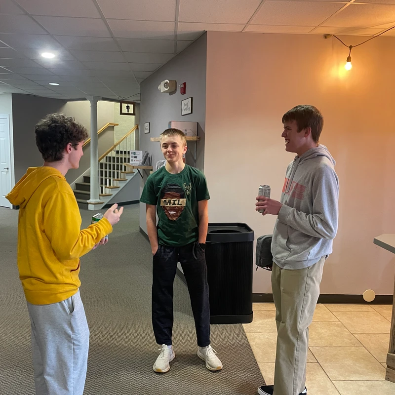 Three young men stand in a circle talking and smiling in a basement common area. One wears a yellow hoodie, one a green t-shirt, and one a grey hoodie.