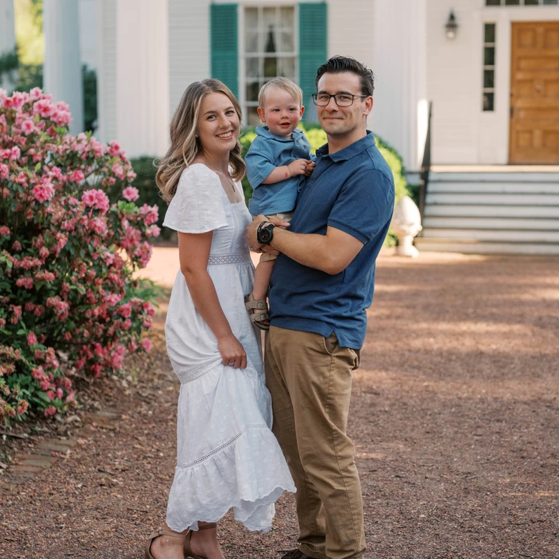 A smiling couple and their baby boy pose for a photo outdoors on a gravel path. The father holds the child. A large white house is in the background.