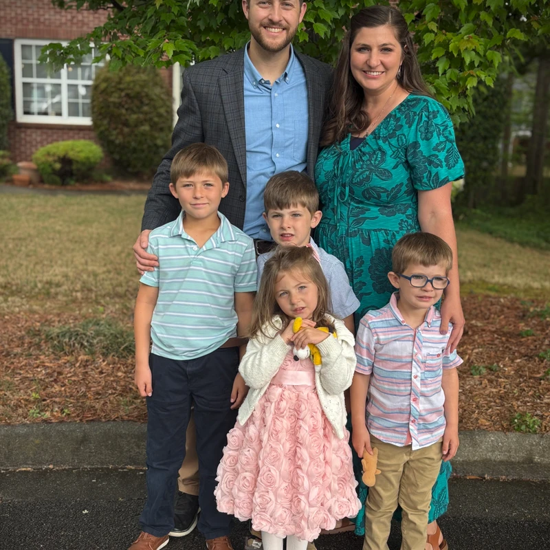 A family of six, including a mother, father, three sons, and a daughter, stand together outdoors smiling for a photo in front of a tree and house.