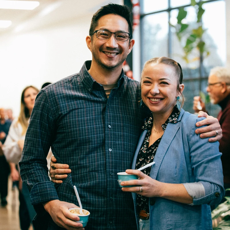 A smiling man and woman pose for a photo at an indoor gathering. The man has his arm around the woman, and they are both holding small cups of dessert.