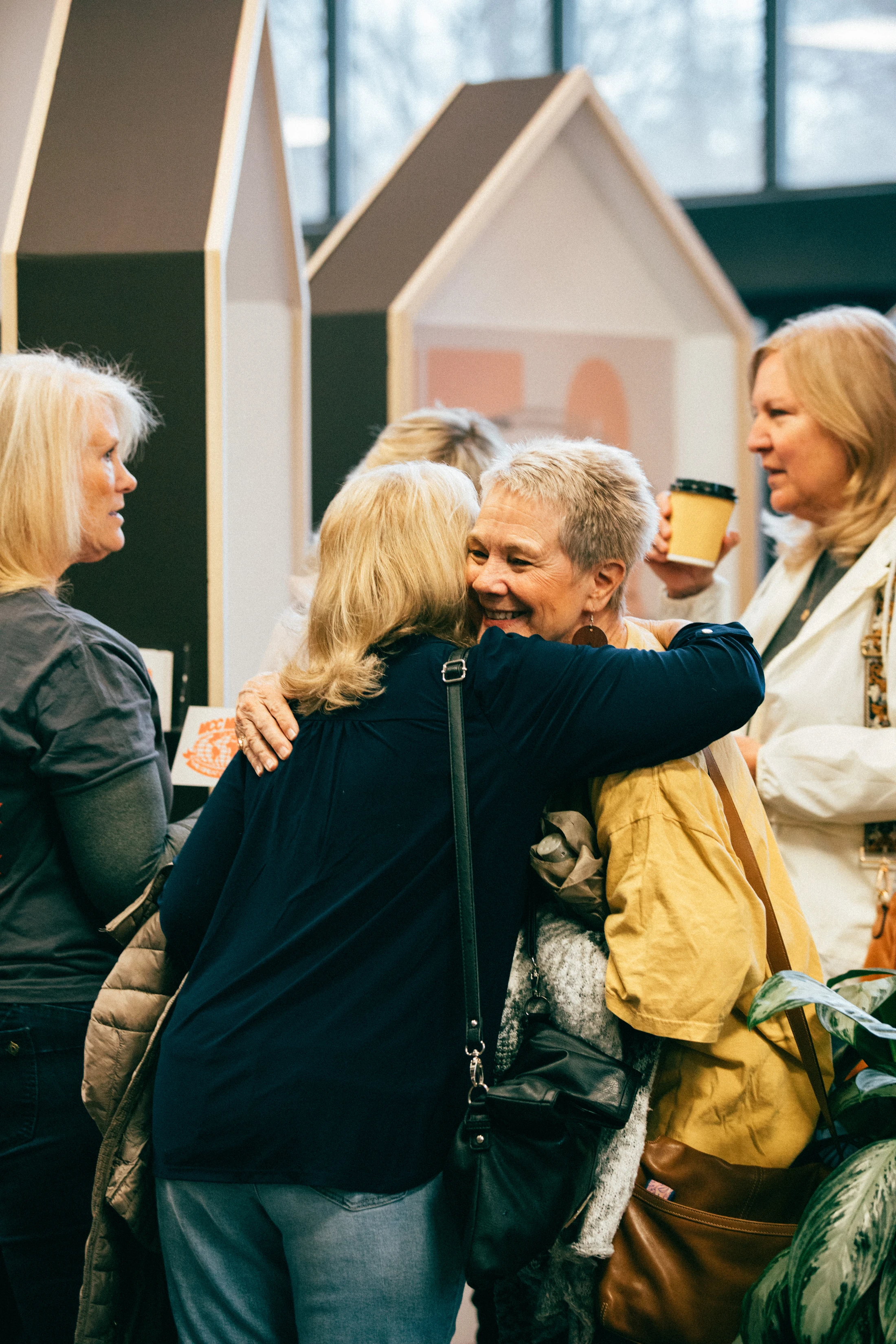Two smiling older women share a happy hug in a crowded room. Other women stand nearby, talking and one holds a coffee cup in a friendly, social setting.