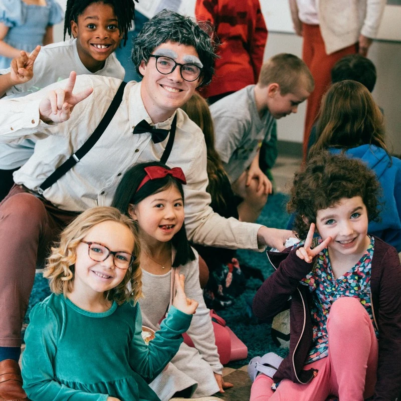 A group of smiling, diverse children pose for a photo with a man wearing a costume of an old man with a grey wig, glasses, and a bow tie.