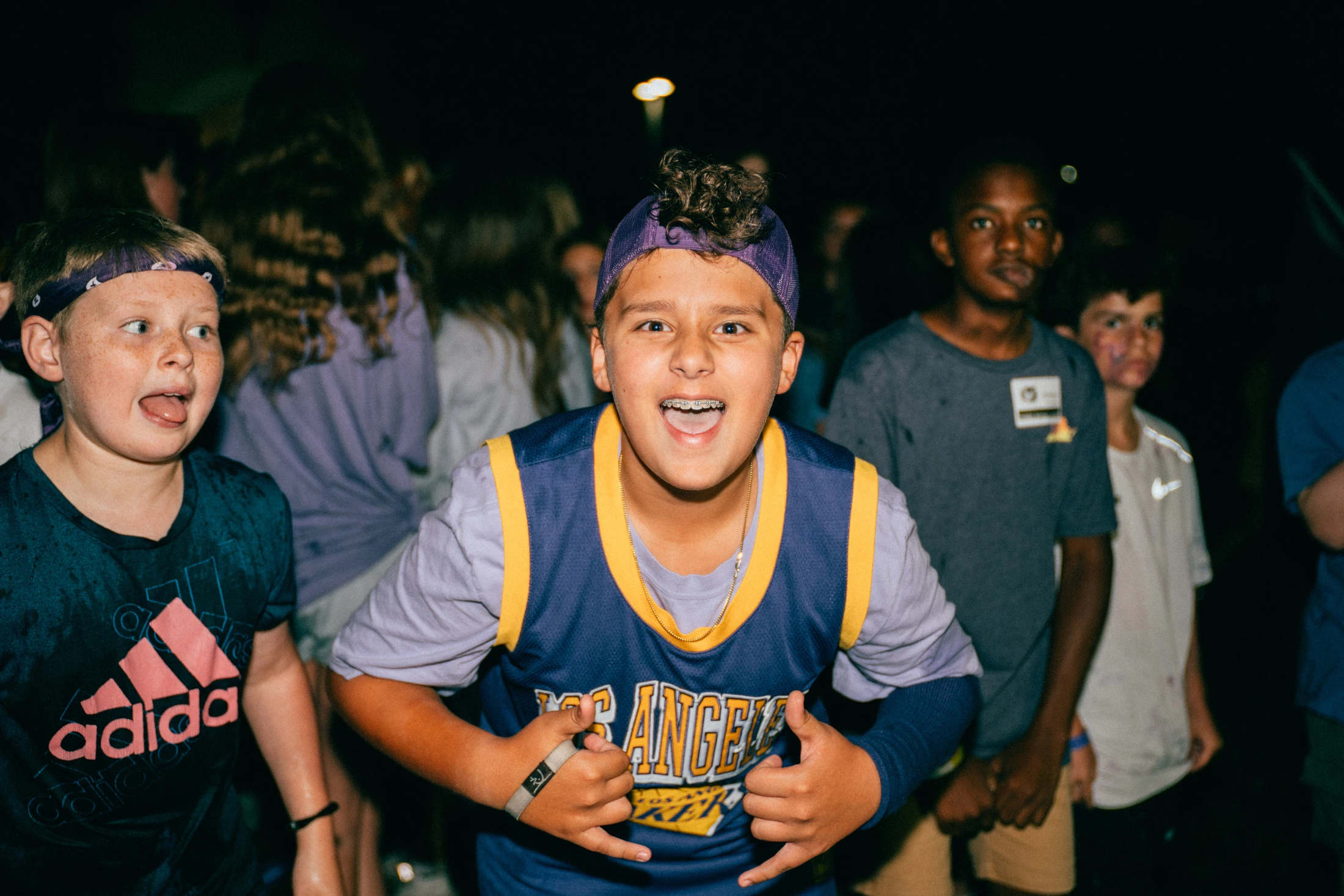 A young boy in a basketball jersey and backward cap smiles with braces, making a hand gesture at the camera, surrounded by other kids at night.