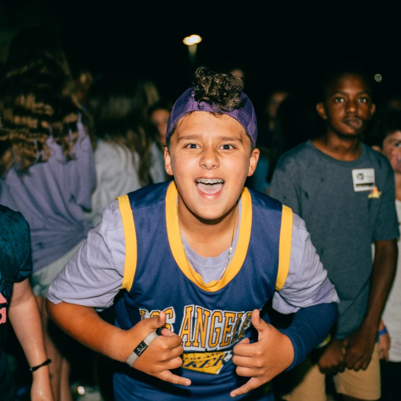 A young boy in a basketball jersey and backward cap smiles with braces, making a hand gesture at the camera, surrounded by other kids at night.