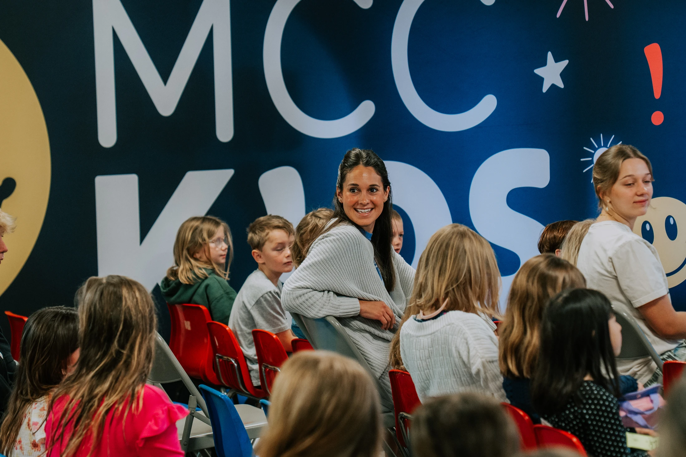 A woman in a grey sweater turns around in her chair to smile at the camera while sitting with a group of children in front of a blue 'MCC KIDS' banner.