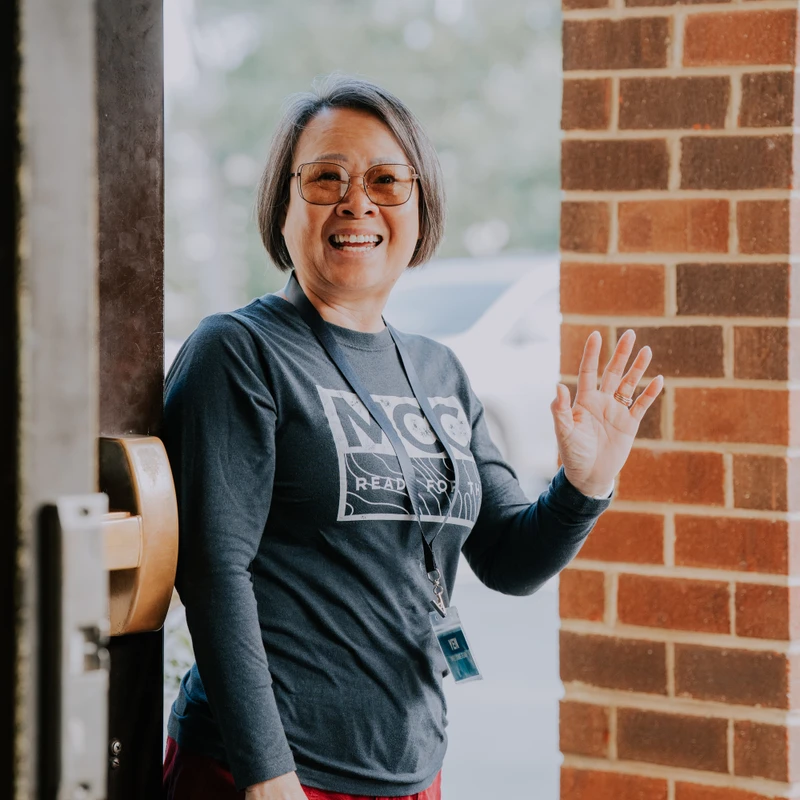 A smiling woman with short hair and glasses stands in a doorway next to a brick wall, waving her hand in a welcoming gesture.