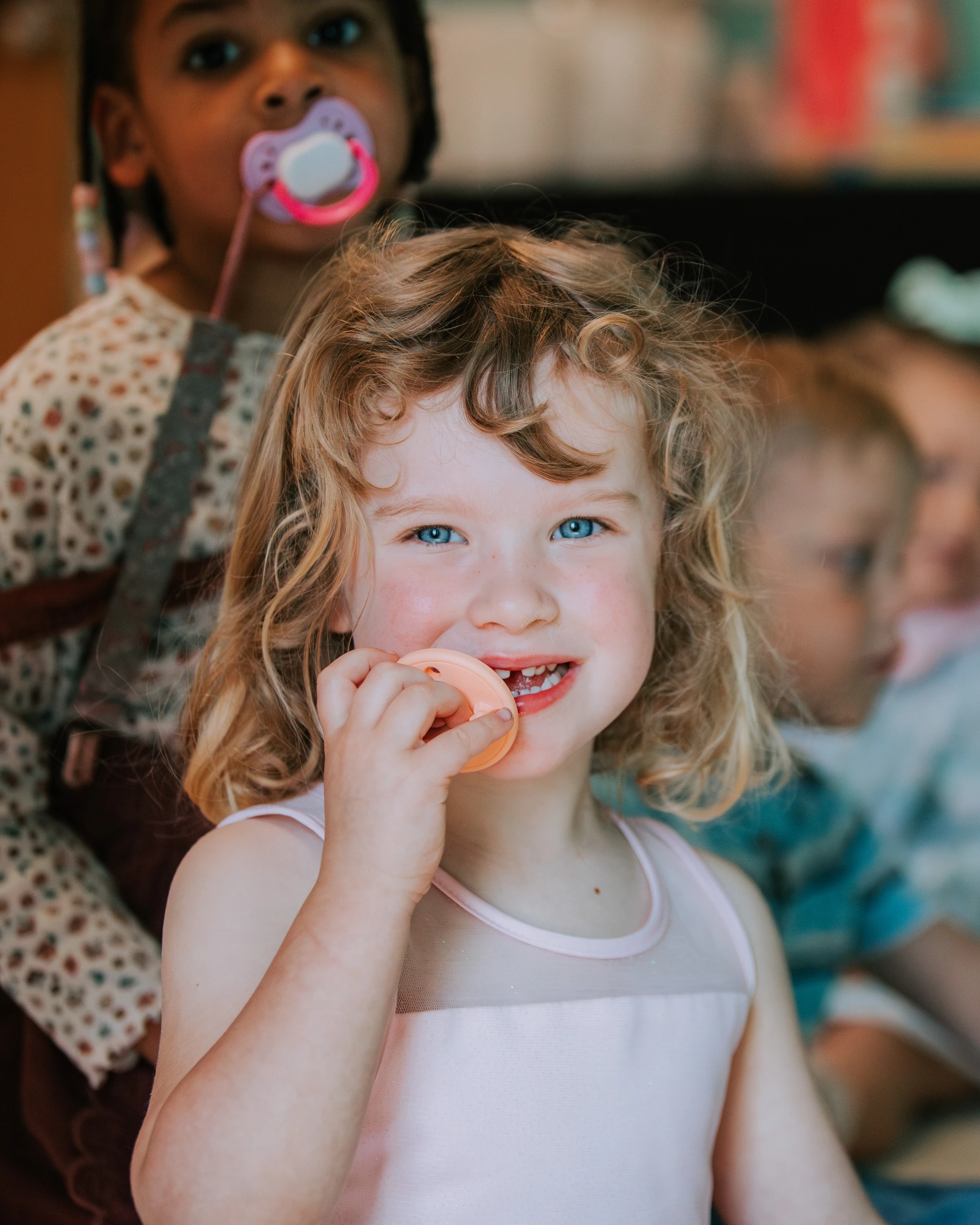 A close-up portrait of a young girl with curly blonde hair and blue eyes. She smiles at the camera while holding a pacifier near her mouth.