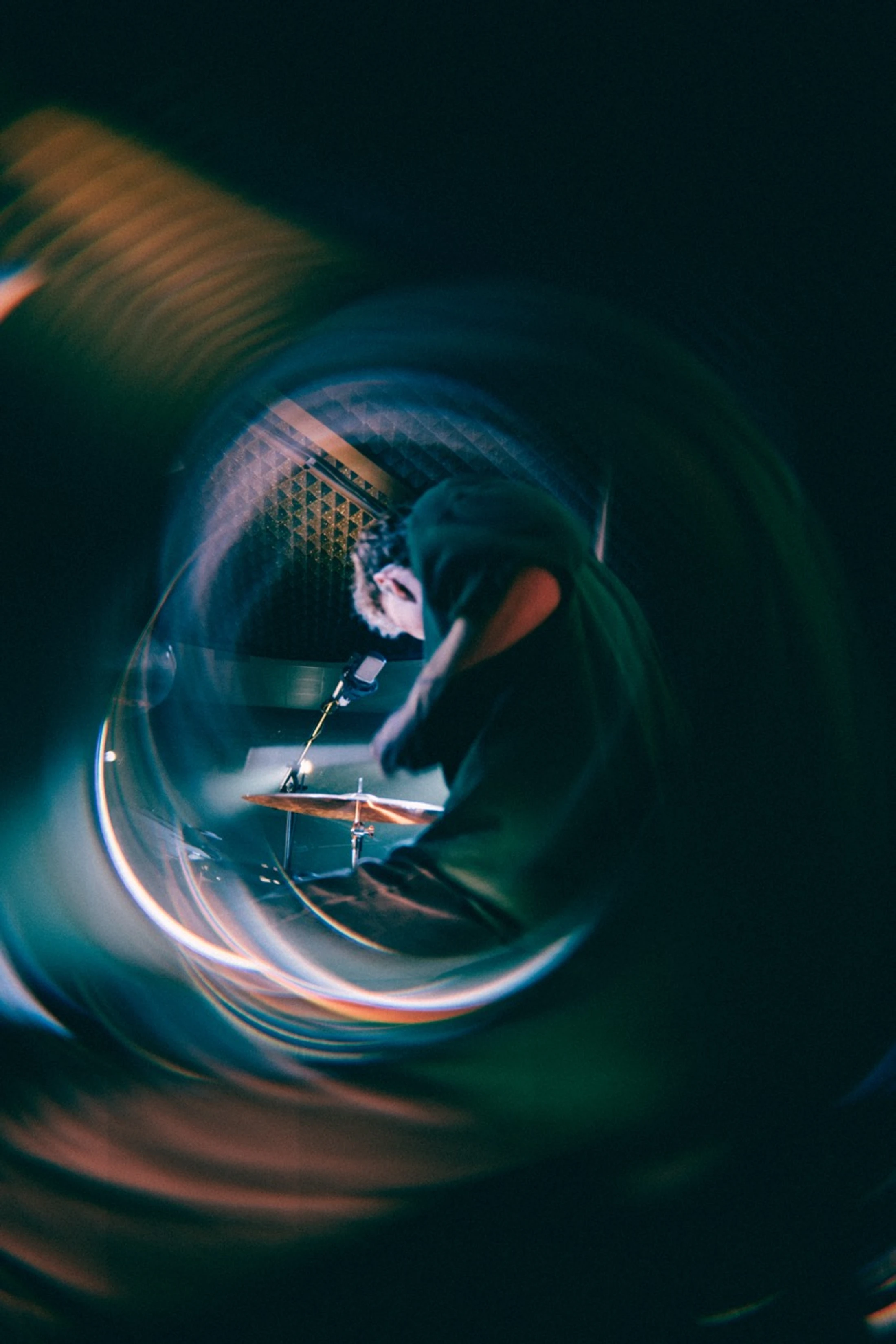 A distorted, swirling view of a drummer in a dark green hoodie playing cymbals in a recording studio, as if seen through a glass or fisheye lens.