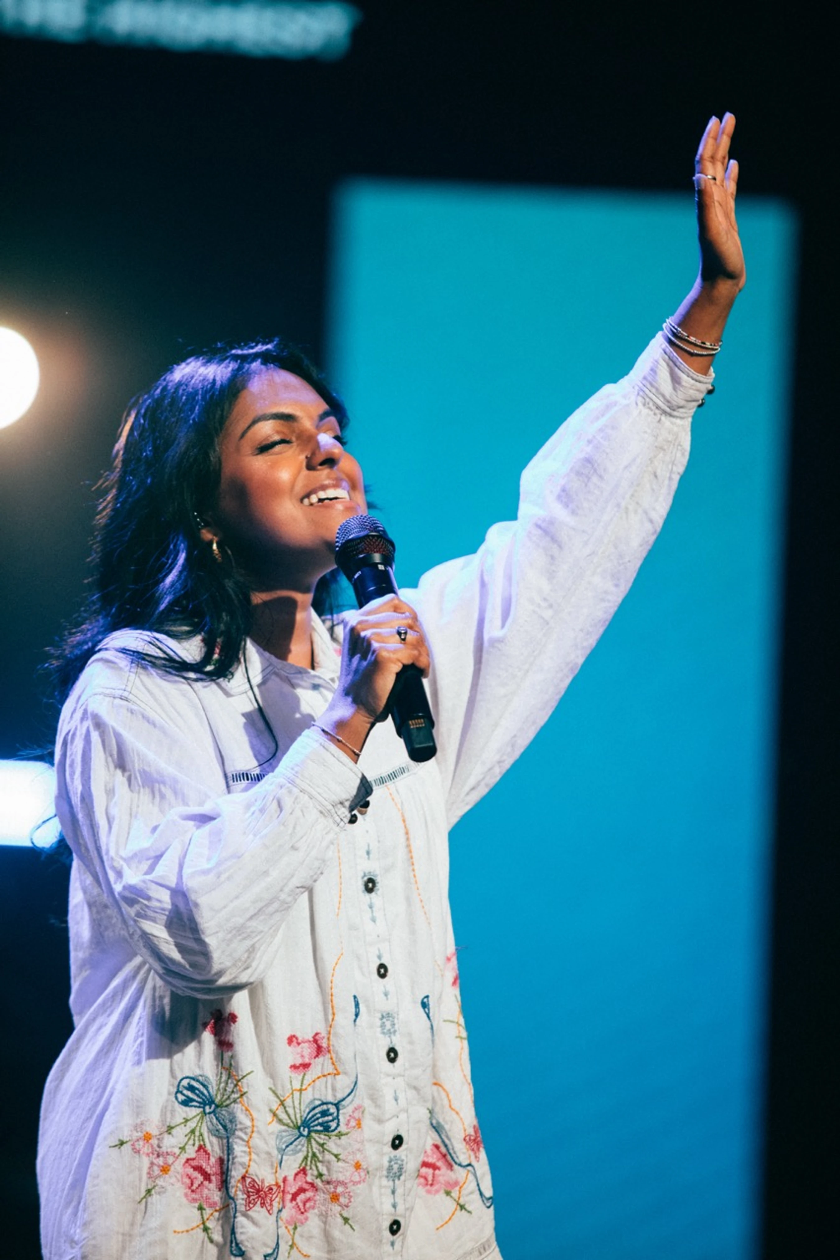 A woman with dark hair sings into a microphone with her eyes closed and one hand raised. She wears a white embroidered shirt on a dimly lit stage.