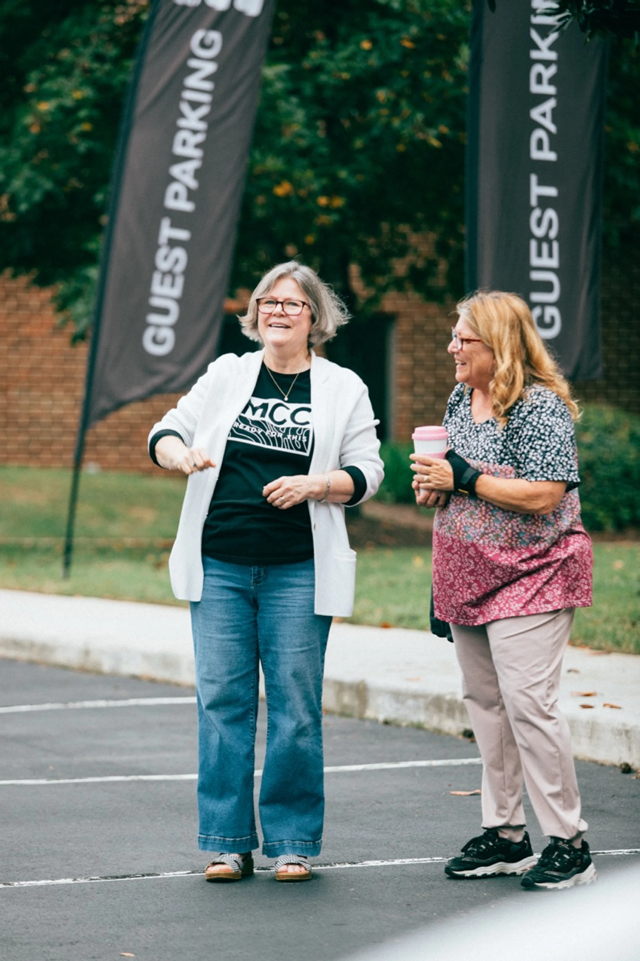 Two older women stand smiling and chatting in a parking lot. In the background, two black banners read "GUEST PARKING" in white text.
