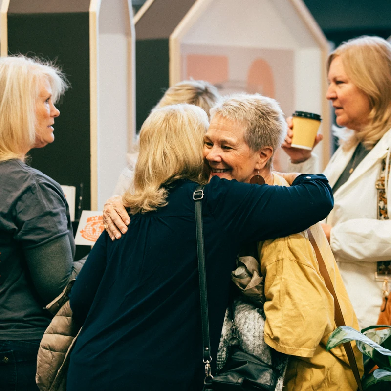 Two women share a joyful hug in the middle of a group. The woman facing the camera has short gray hair and a big smile. Other women look on and chat.