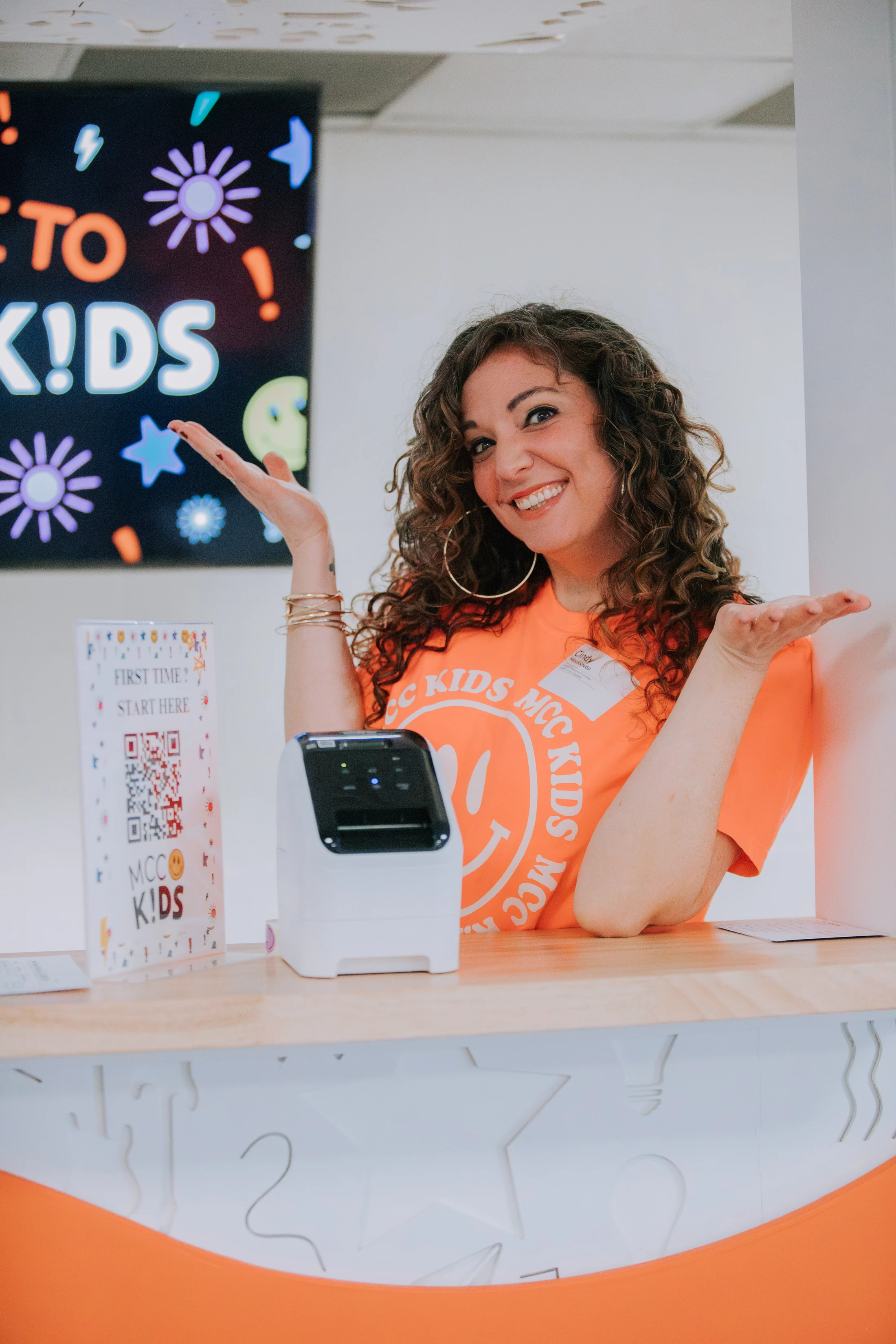 A smiling woman with curly hair in an orange t-shirt welcomes people from behind a check-in desk with a label printer and a QR code sign.