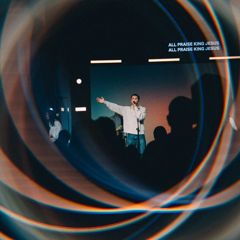 A man on a dimly lit stage sings into a microphone, with lyrics on a screen behind him, viewed through a swirling, colorful light vortex effect.