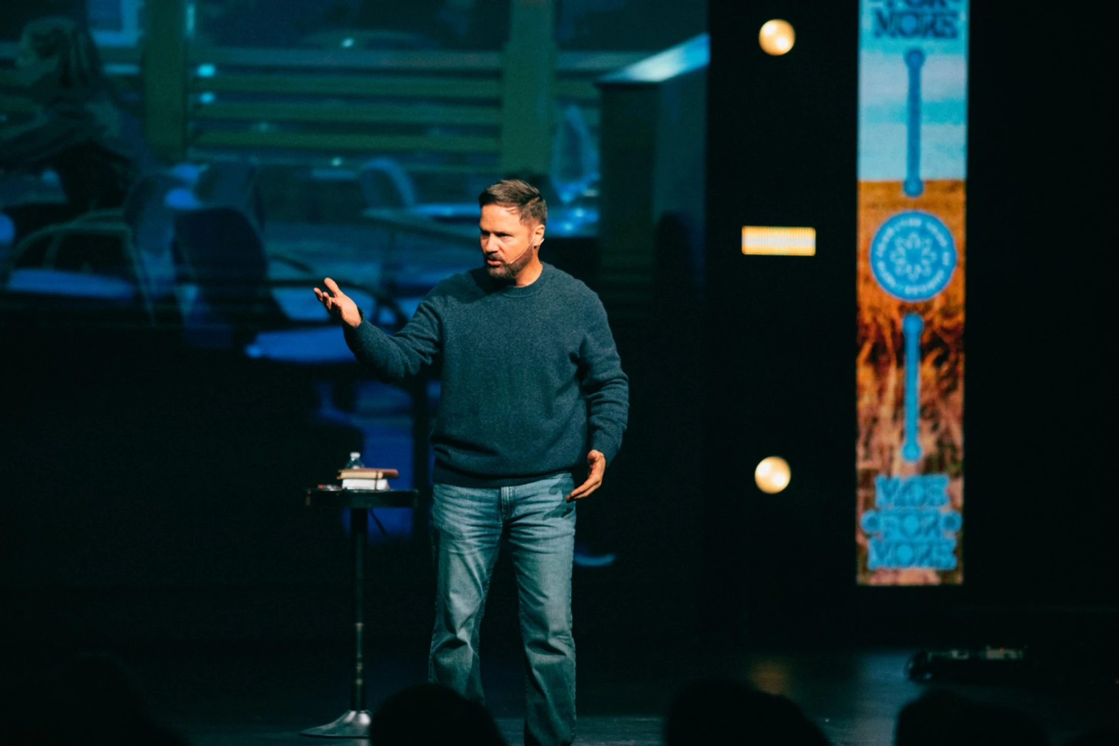 A man in a blue sweater and jeans stands on a dimly lit stage next to a small table, gesturing with his hand as he speaks to an audience.