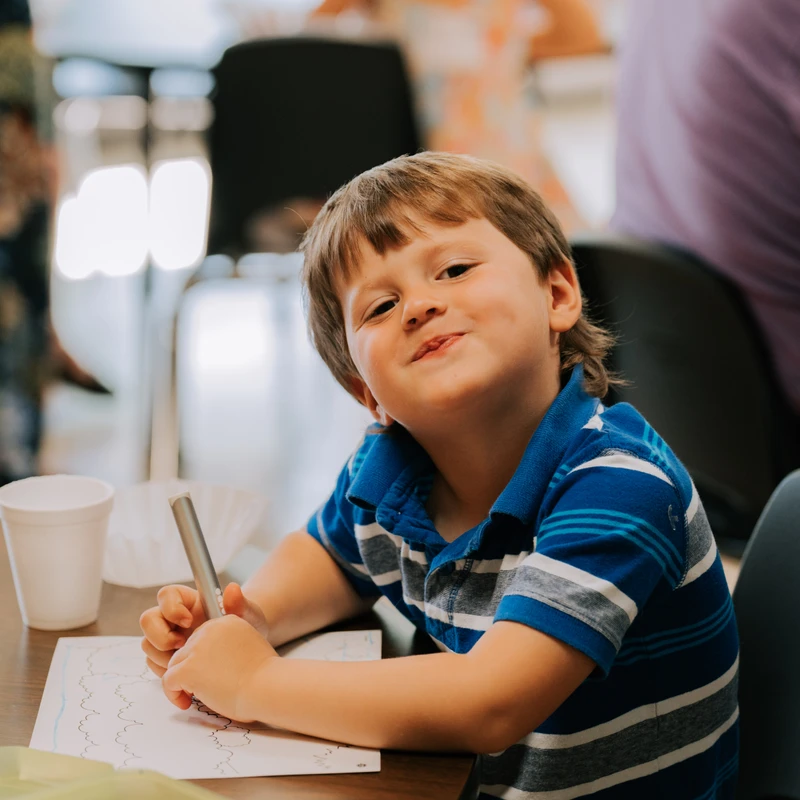 A young boy in a blue striped polo shirt sits at a table, holding a pen over a piece of paper. He looks up at the camera with a happy, playful expression.
