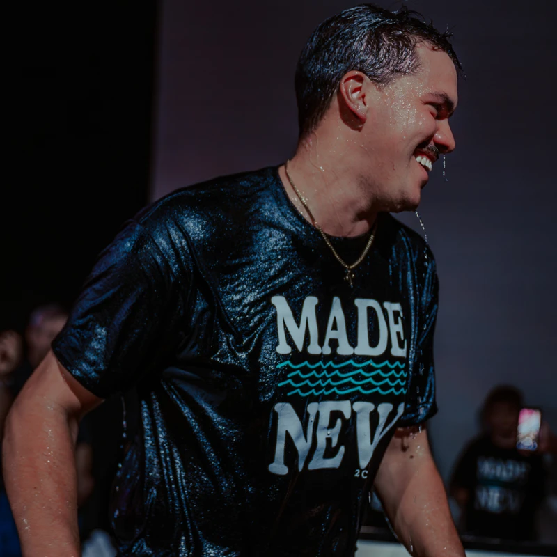 A smiling young man, soaking wet, stands in profile after his baptism. He wears a black t-shirt that says 'MADE NEW' as water drips from his face and hair.
