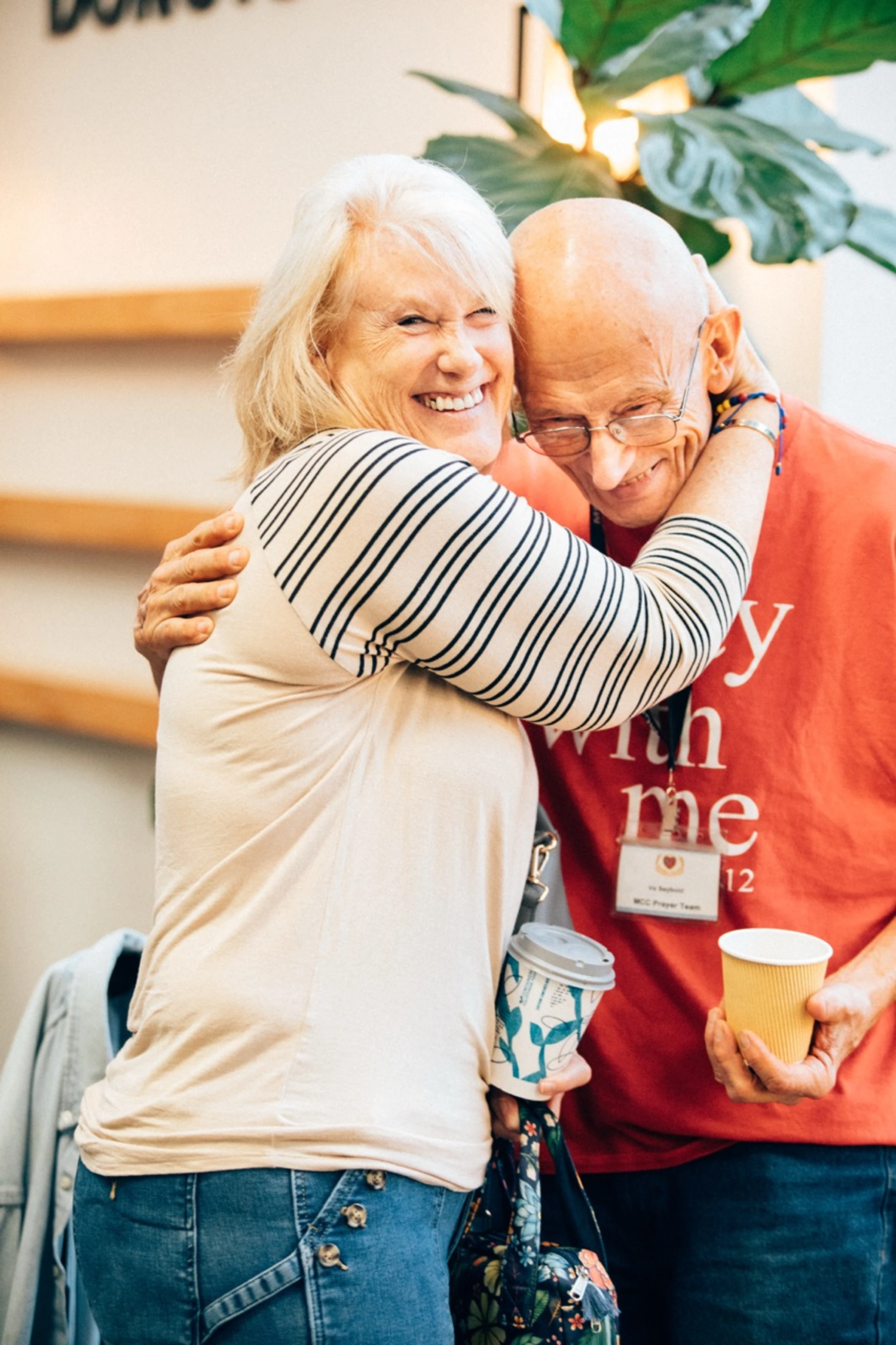 A smiling older woman with blonde hair hugs an older bald man with glasses. They both look happy and are holding coffee cups at an indoor event.