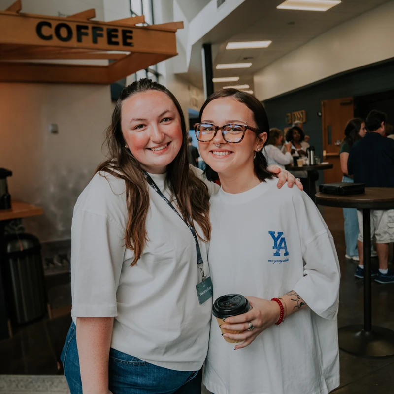 Two young women stand together smiling, with one's arm around the other. They are indoors in front of a coffee bar with other people in the background.