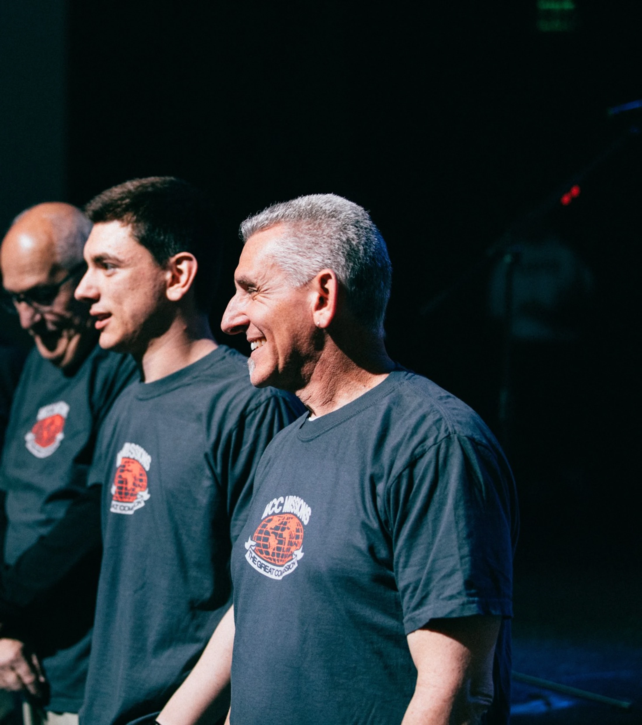 Three men of varying ages stand together in profile, smiling. They all wear matching gray t-shirts with a red logo for 'ACC Missions The Great Commission'.