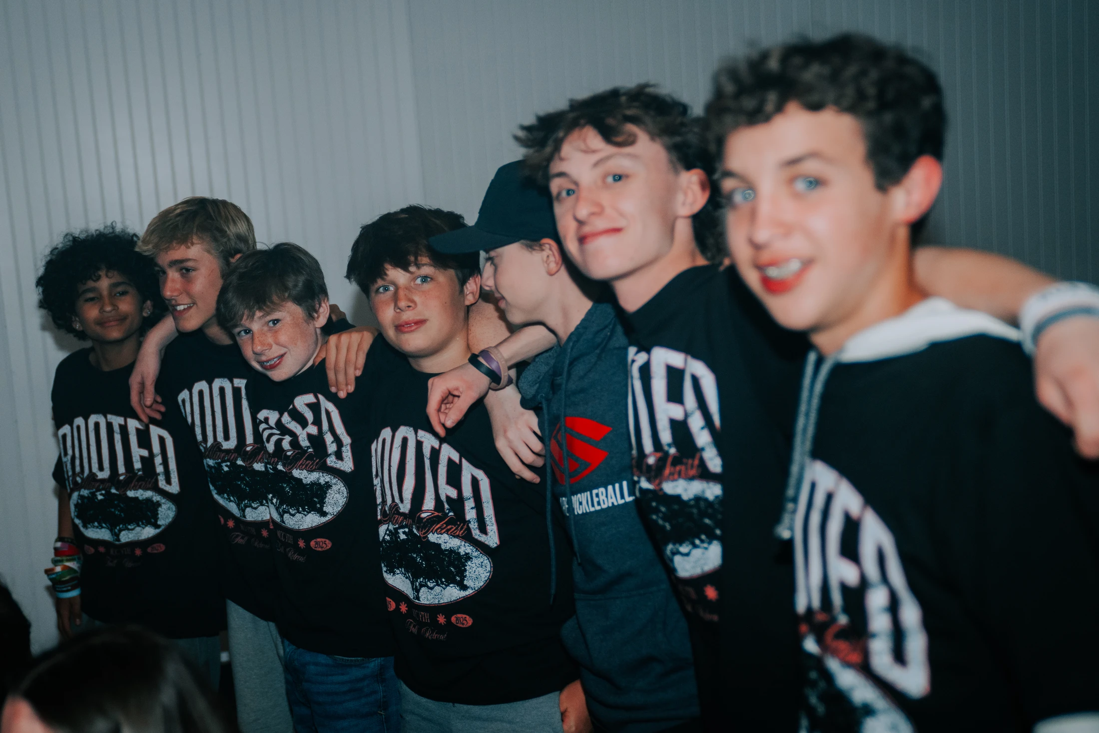 A line of teenage boys with their arms around each other's shoulders, smiling at the camera. Most are wearing matching black 'ROOTED' t-shirts.
