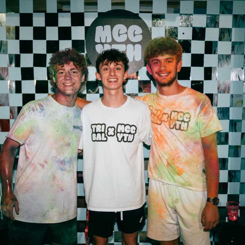 Three young men smile for a photo, two in paint-splattered tie-dye shirts, in front of a checkered wall with graffiti at what appears to be a youth event.