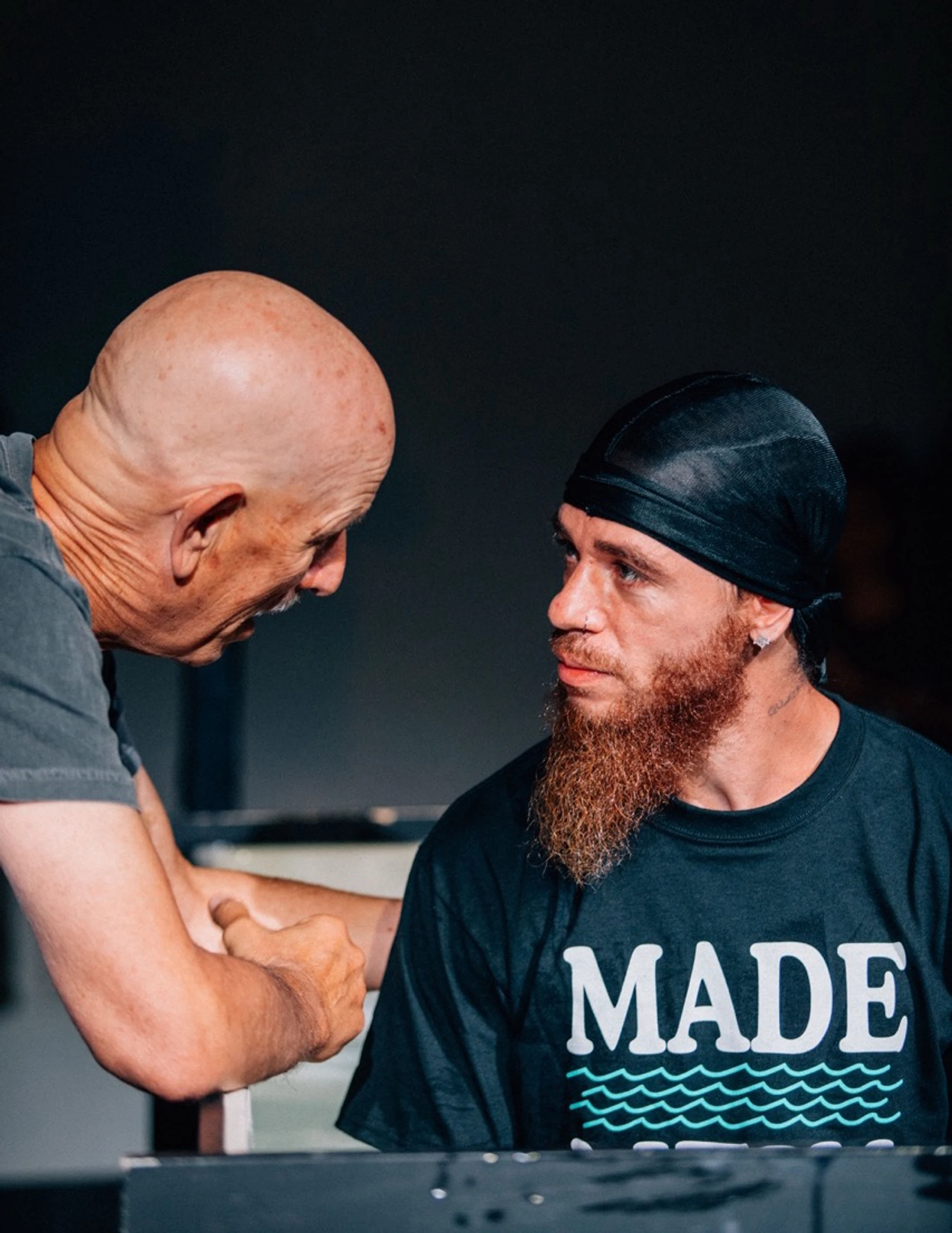 An older, bald man in a grey shirt leans in to speak with a younger man with a red beard wearing a black durag and a t-shirt that says 'MADE'.