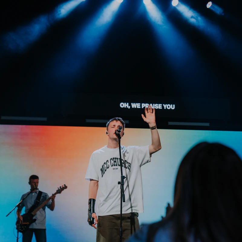 A young man sings into a microphone on a dimly lit stage, his hand raised. Behind him, lyrics on a screen read 'OH, WE PRAISE YOU' under blue spotlights.