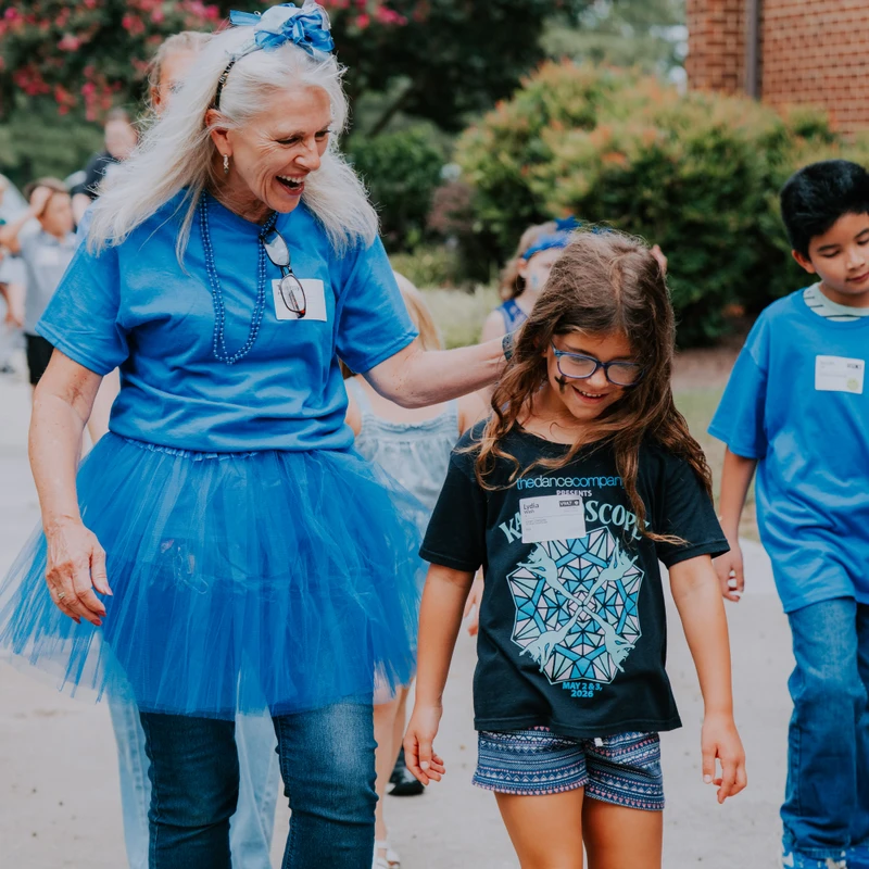 An older woman in a blue t-shirt and tutu smiles while walking with her arm on a young girl's shoulder during an outdoor event with other children.