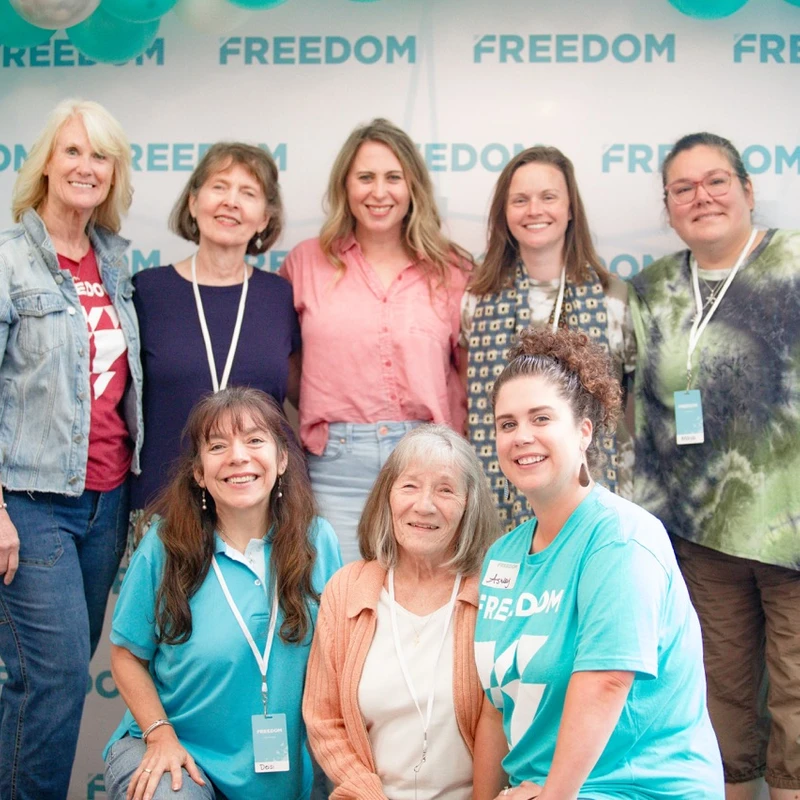 A group of eight women smile for a photo in front of a backdrop with the word 'FREEDOM' and teal, white, and silver balloons.