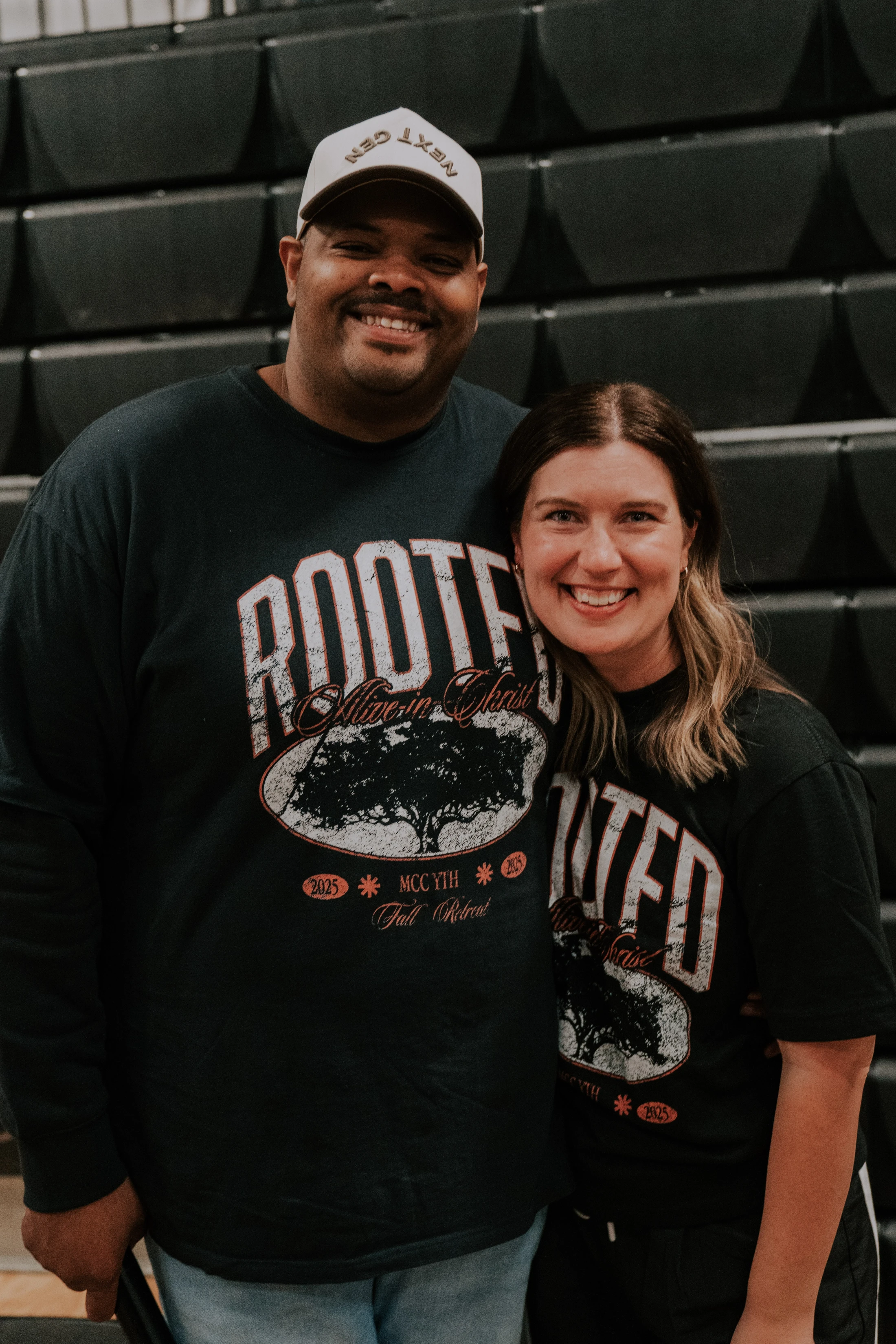 A smiling man and woman stand together for a photo, wearing matching black t-shirts for an MCC YTH Fall Retreat event against a dark background.