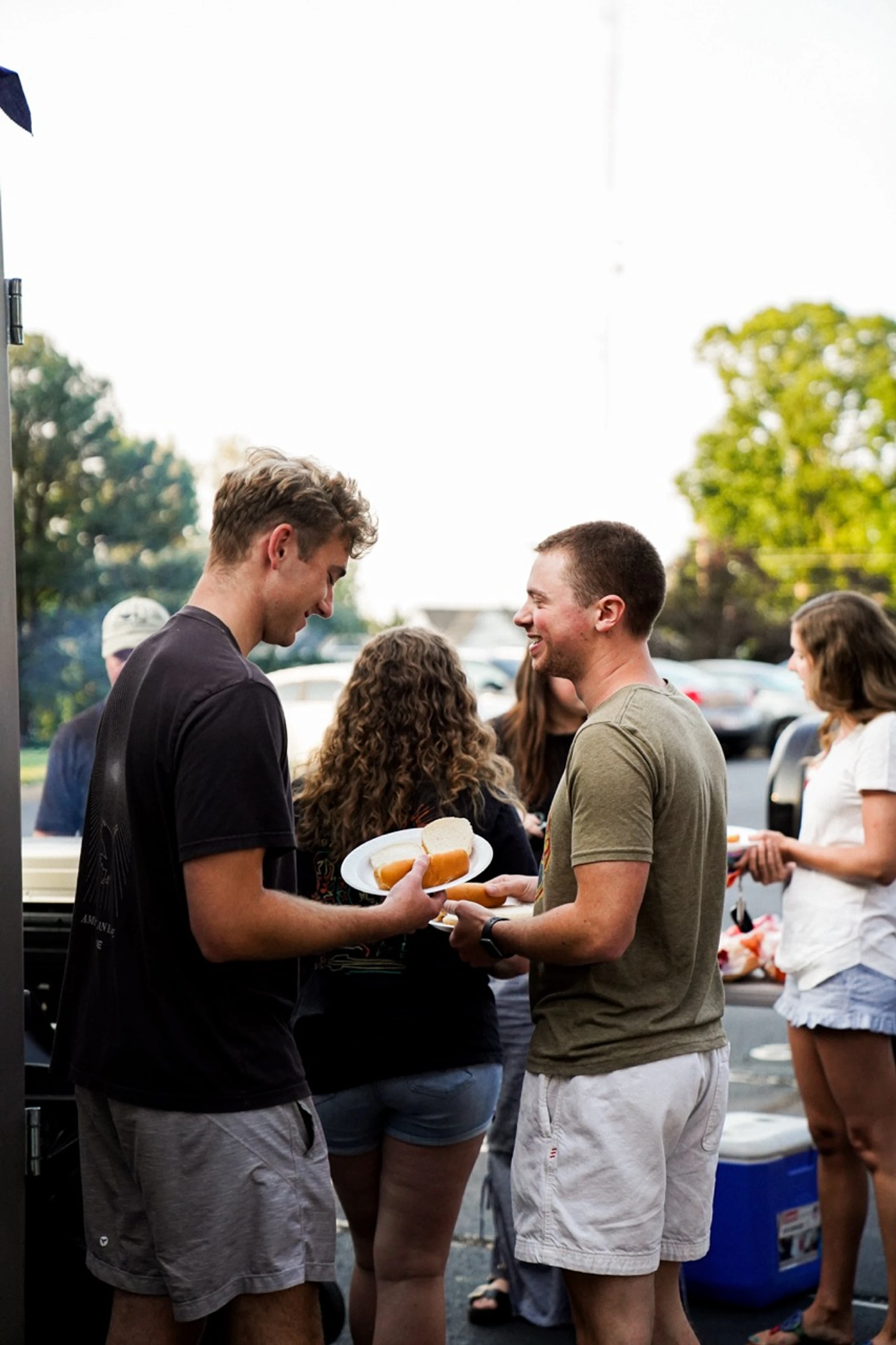 Two young men smile as one hands the other a plate with a hot dog in a bun. Other people are gathered around at an outdoor cookout in a parking lot.