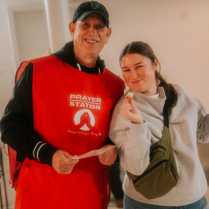 An older man in a red 'Prayer Station' apron and a young woman in a grey sweatshirt smile and pose together for a photo indoors.
