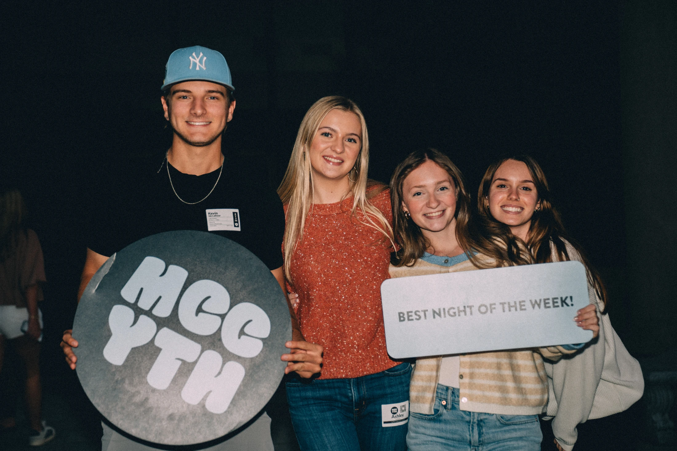 Four smiling young adults pose together at an MCC Youth event, holding signs that say 'MCC YTH' and 'Best Night of the Week!'.