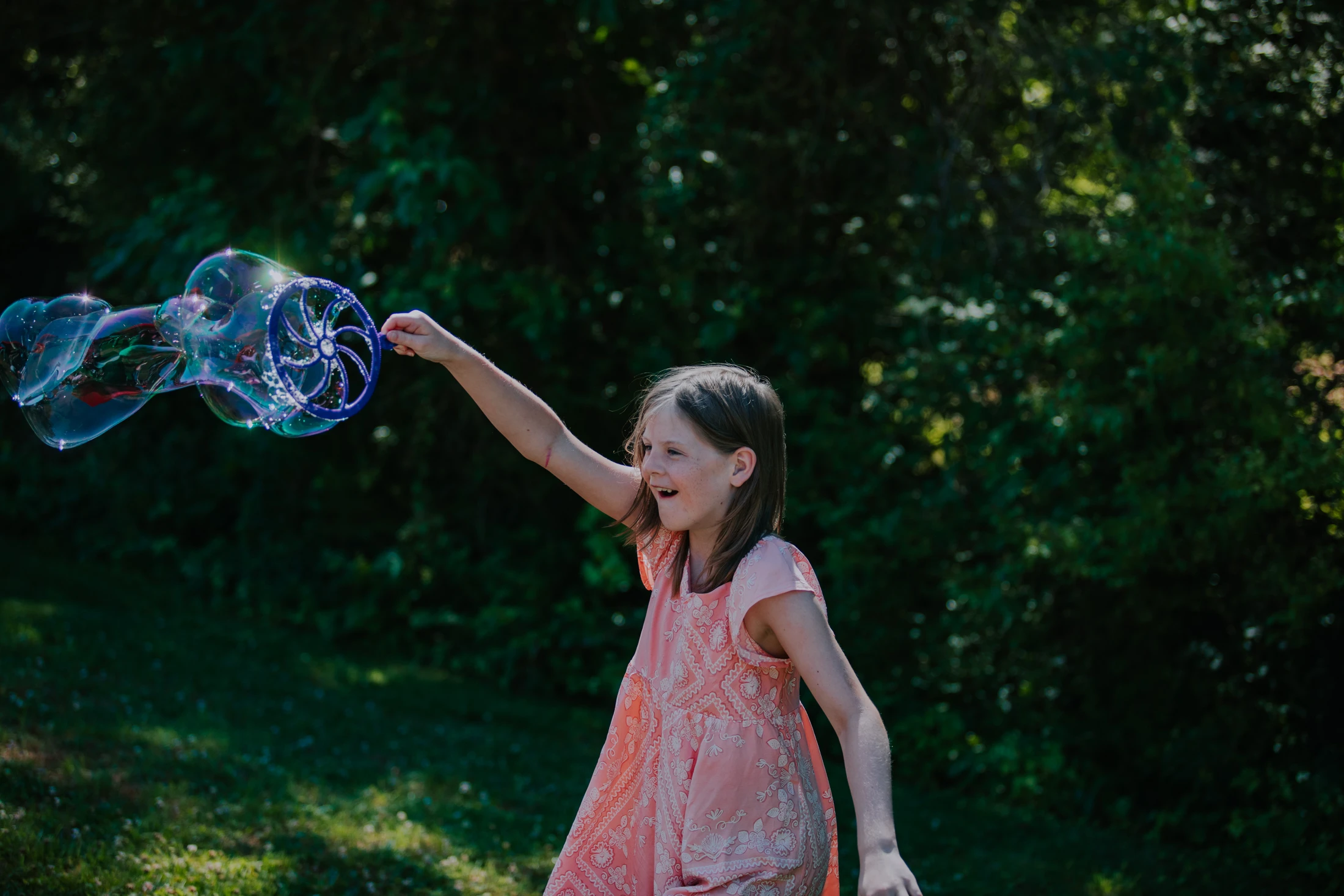 A young girl in a pink dress smiles happily as she creates giant, shimmering soap bubbles with a large blue wand in a sunlit grassy area.