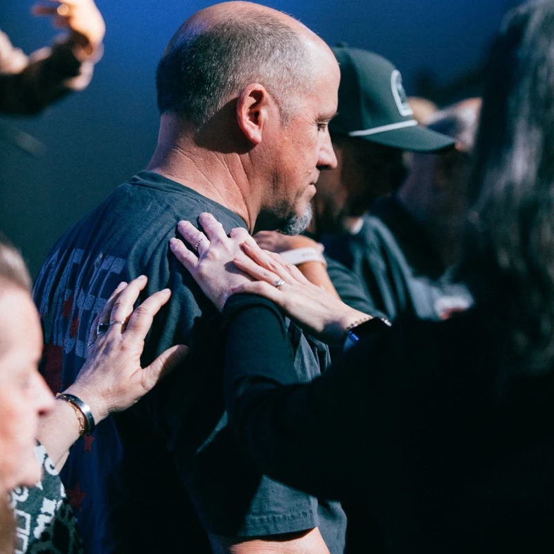 A balding man in profile has several hands resting on his shoulders in a comforting gesture during what appears to be a religious service or group prayer.